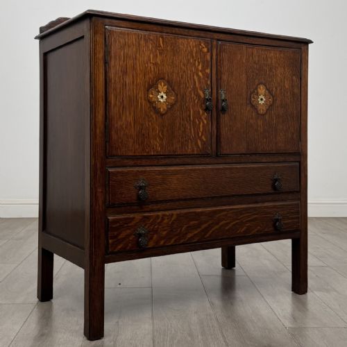 fig 7. A dark oak Arts and Crafts cupboard featuring double panelled doors with floral marquetry inlays above two wide drawers, finished with dark metal Celtic knot handles.