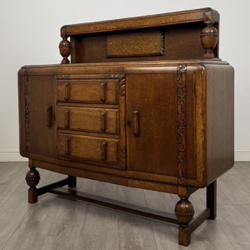 fig 1. A medium brown oak Jacobean style sideboard featuring a raised gallery shelf, carved side columns, three central drawers, flanking cupboard doors, and bulbous turned legs connected by a stretcher.