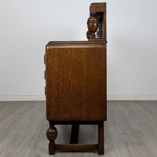 fig 2. A medium brown oak Jacobean style sideboard featuring a raised gallery shelf, carved side columns, three central drawers, flanking cupboard doors, and bulbous turned legs connected by a stretcher.