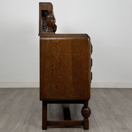 fig 6. A medium brown oak Jacobean style sideboard featuring a raised gallery shelf, carved side columns, three central drawers, flanking cupboard doors, and bulbous turned legs connected by a stretcher.