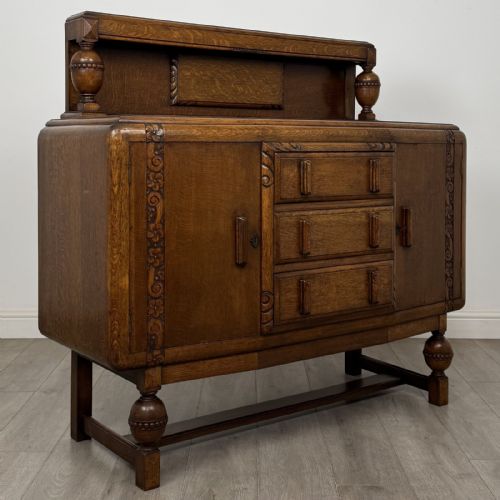 fig 7. A medium brown oak Jacobean style sideboard featuring a raised gallery shelf, carved side columns, three central drawers, flanking cupboard doors, and bulbous turned legs connected by a stretcher.