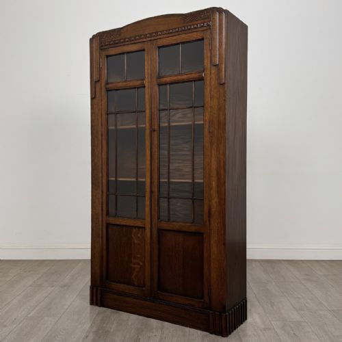 fig 1. A tall dark oak Art Deco bookcase featuring two glazed doors with geometric glazing bars, stylised corner carvings, and a subtle arched top frieze.