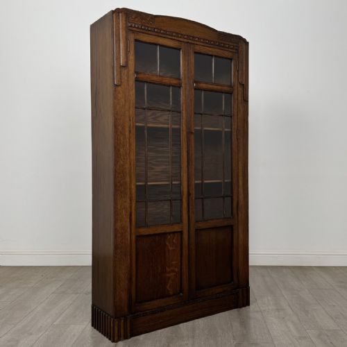 fig 7. A tall dark oak Art Deco bookcase featuring two glazed doors with geometric glazing bars, stylised corner carvings, and a subtle arched top frieze.