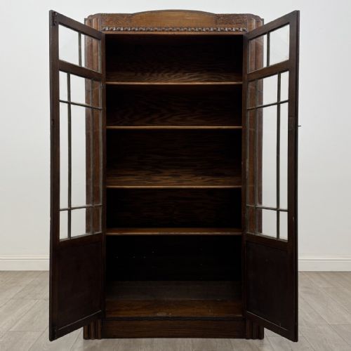 fig 8. A tall dark oak Art Deco bookcase featuring two glazed doors with geometric glazing bars, stylised corner carvings, and a subtle arched top frieze.