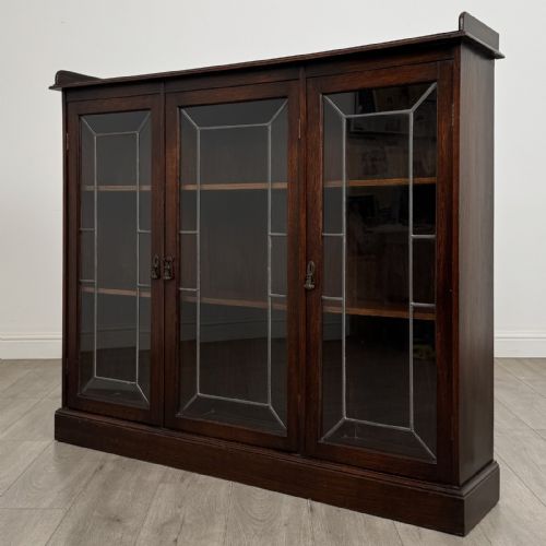 fig 2. An Edwardian dark oak display cabinet featuring three doors with geometric leaded glass panels and interior wooden shelves.