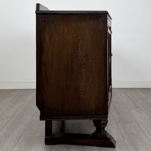 fig 7. A dark brown Jacobean style oak sideboard featuring three central drawers flanked by two geometric panel cupboard doors, supported on turned legs.