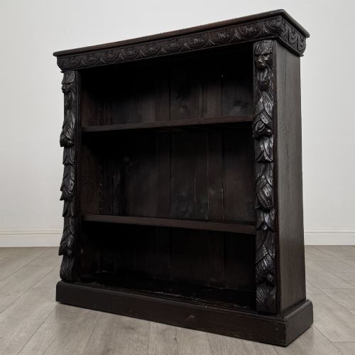 fig 2. A dark carved oak open bookcase featuring three shelf surfaces and pilasters detailed with foliate carvings and Green Man masks.