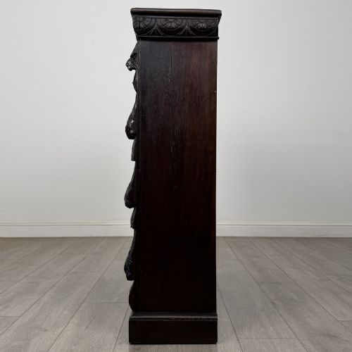 fig 3. A dark carved oak open bookcase featuring three shelf surfaces and pilasters detailed with foliate carvings and Green Man masks.