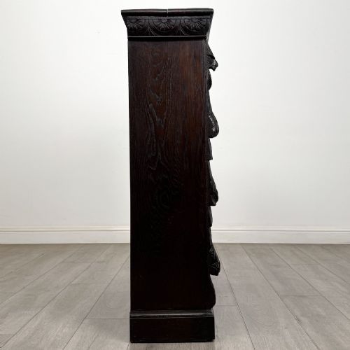 fig 7. A dark carved oak open bookcase featuring three shelf surfaces and pilasters detailed with foliate carvings and Green Man masks.