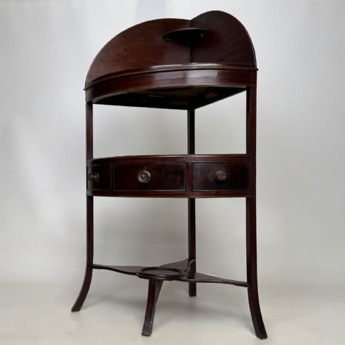 fig 1. A dark mahogany Georgian bow-front corner washstand featuring a curved splashback with a small upper shelf, a middle section with a central drawer flanked by dummy drawers, and splayed legs united by a circular stretcher.