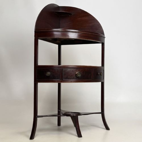fig 3. A dark mahogany Georgian bow-front corner washstand featuring a curved splashback with a small upper shelf, a middle section with a central drawer flanked by dummy drawers, and splayed legs united by a circular stretcher.