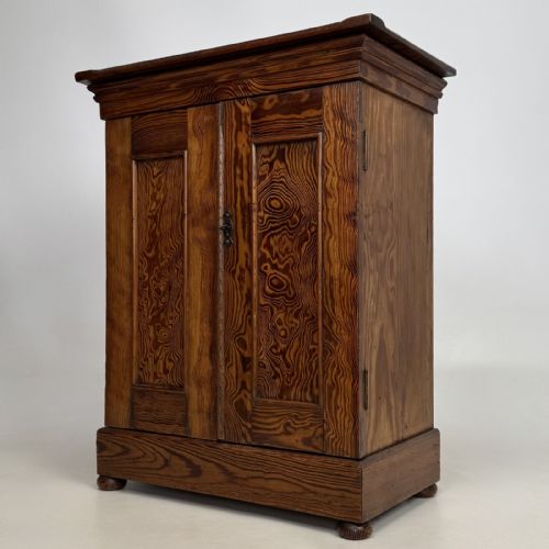 fig 1. A small Victorian pitch pine cupboard featuring twin panelled doors with pronounced wood grain, a moulded top, and a plinth base on small bun feet.