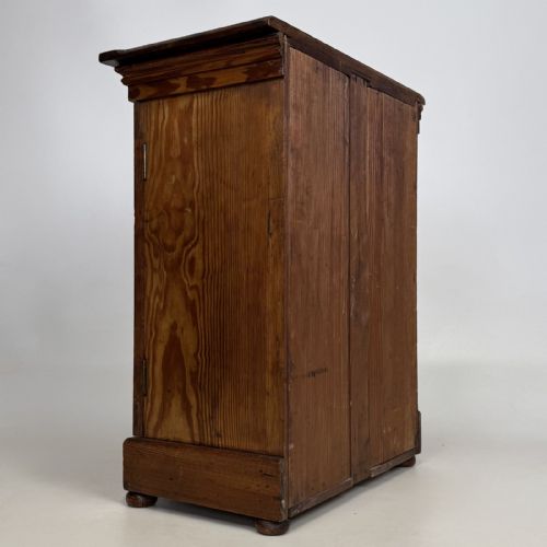 fig 3. A small Victorian pitch pine cupboard featuring twin panelled doors with pronounced wood grain, a moulded top, and a plinth base on small bun feet.