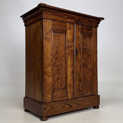 fig 7. A small Victorian pitch pine cupboard featuring twin panelled doors with pronounced wood grain, a moulded top, and a plinth base on small bun feet.