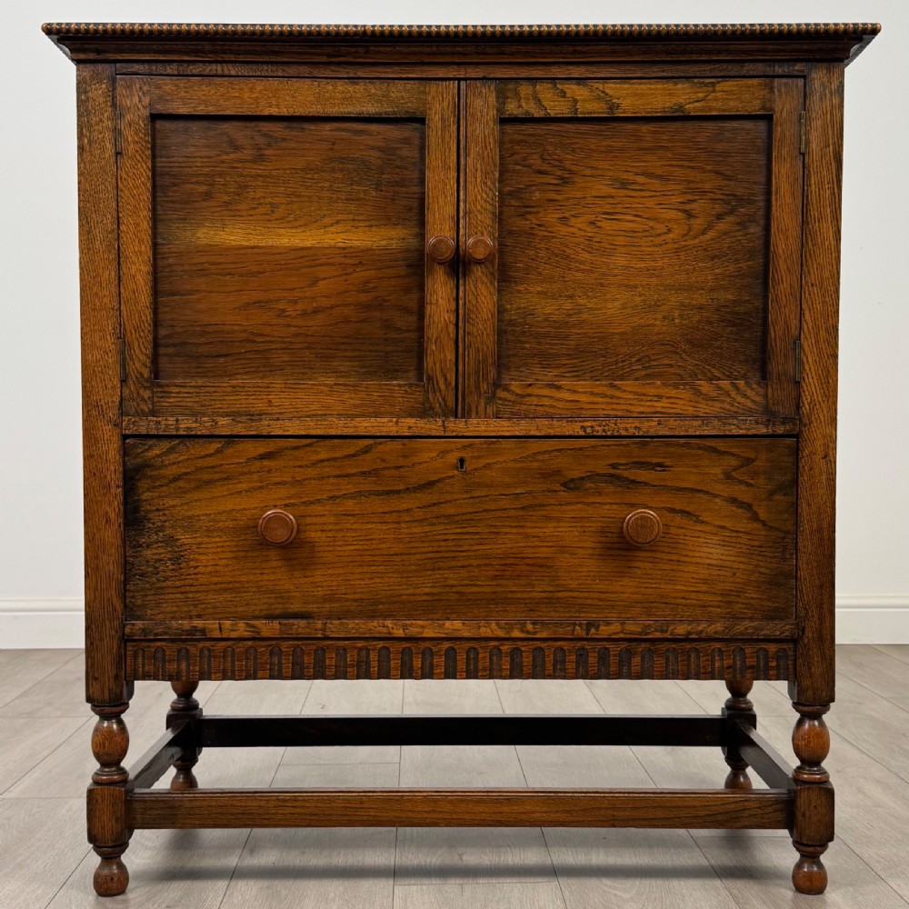 An antique twentieth-century golden oak cupboard featuring two panelled upper doors, a single lower drawer, and raised turned supports with an undertier shelf.