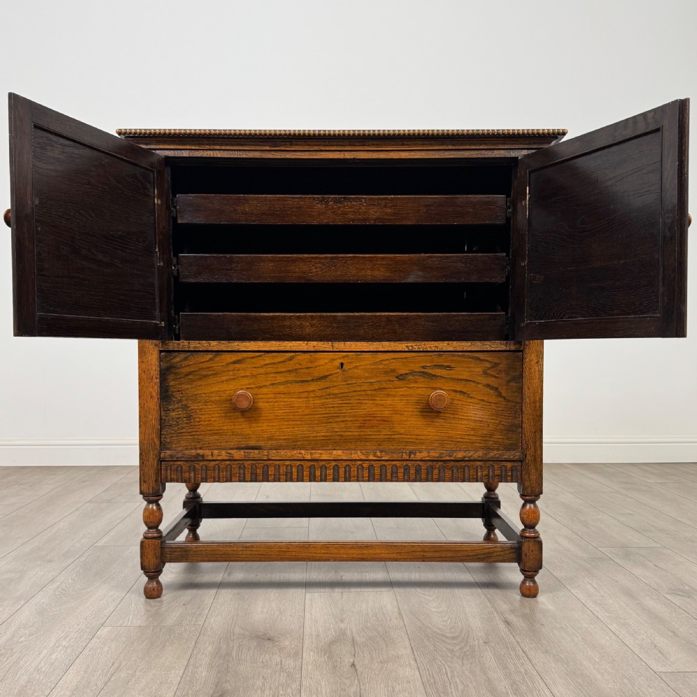 An antique twentieth-century golden oak cupboard featuring two panelled upper doors, a single lower drawer, and raised turned supports with an undertier shelf. View II