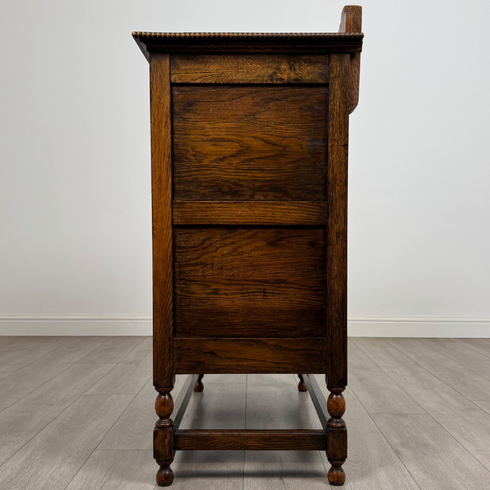 An antique twentieth-century golden oak cupboard featuring two panelled upper doors, a single lower drawer, and raised turned supports with an undertier shelf. View IV