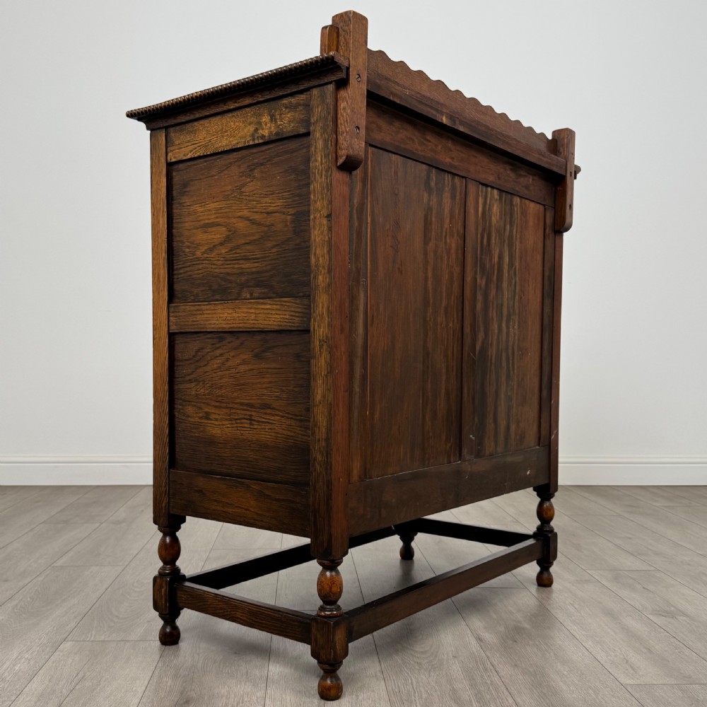 An antique twentieth-century golden oak cupboard featuring two panelled upper doors, a single lower drawer, and raised turned supports with an undertier shelf. View VI