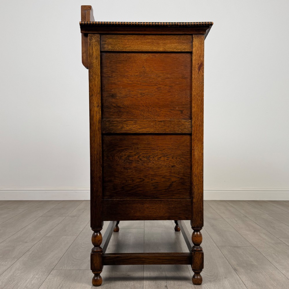 An antique twentieth-century golden oak cupboard featuring two panelled upper doors, a single lower drawer, and raised turned supports with an undertier shelf. View IX