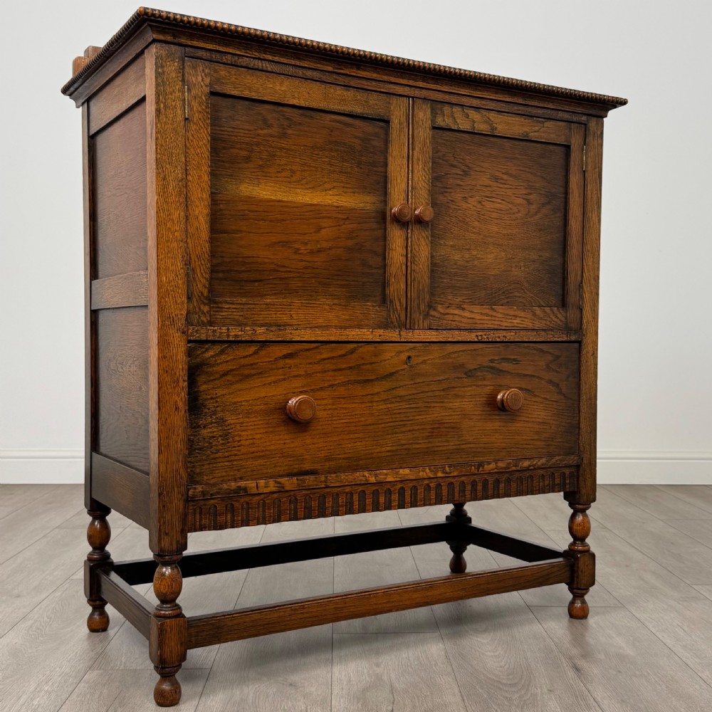 An antique twentieth-century golden oak cupboard featuring two panelled upper doors, a single lower drawer, and raised turned supports with an undertier shelf. View X