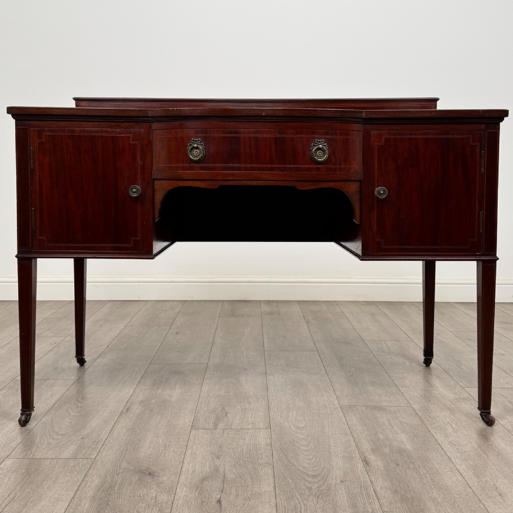 An Edwardian mahogany writing table featuring a central drawer, flanking cupboard doors, boxwood inlay, brass ring handles, and tapered legs on castors.