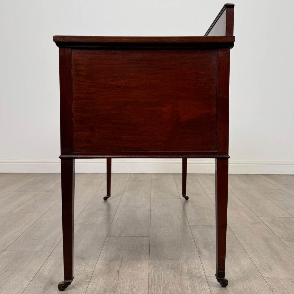 An Edwardian mahogany writing table featuring a central drawer, flanking cupboard doors, boxwood inlay, brass ring handles, and tapered legs on castors. View IV