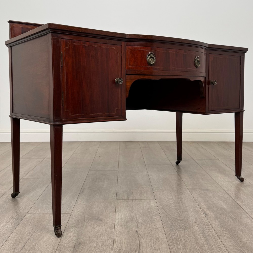 An Edwardian mahogany writing table featuring a central drawer, flanking cupboard doors, boxwood inlay, brass ring handles, and tapered legs on castors. View IX