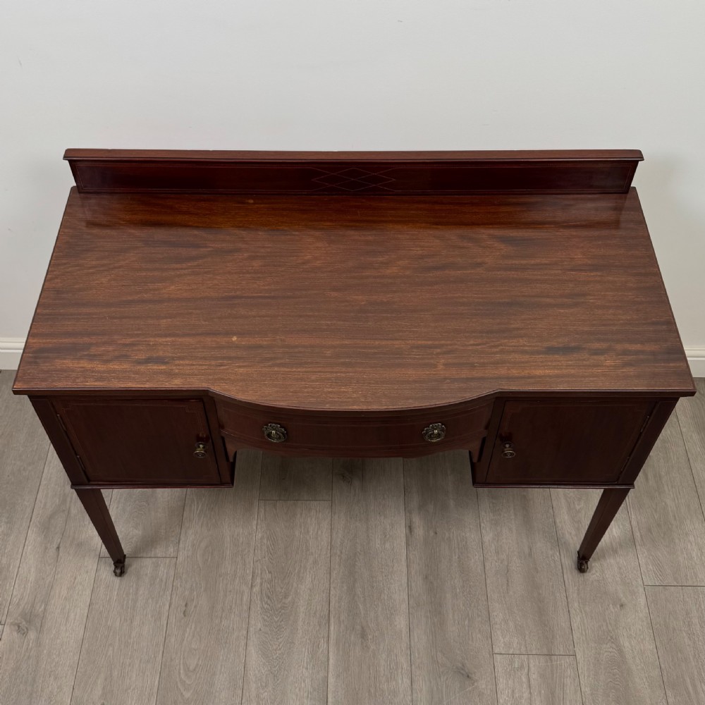 An Edwardian mahogany writing table featuring a central drawer, flanking cupboard doors, boxwood inlay, brass ring handles, and tapered legs on castors. View X
