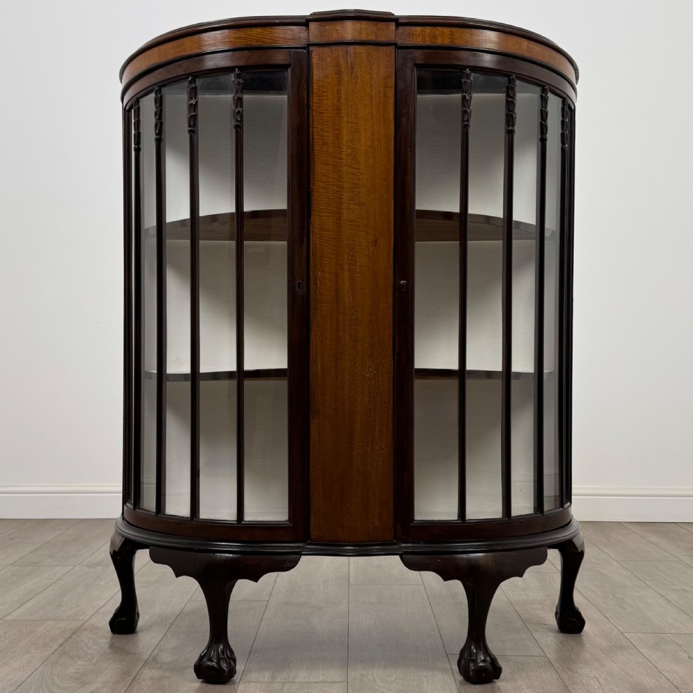 An early twentieth-century mahogany bow-front display cabinet featuring curved glass doors, vertical glazing bars, internal shelves, and cabriole legs with ball-and-claw feet.