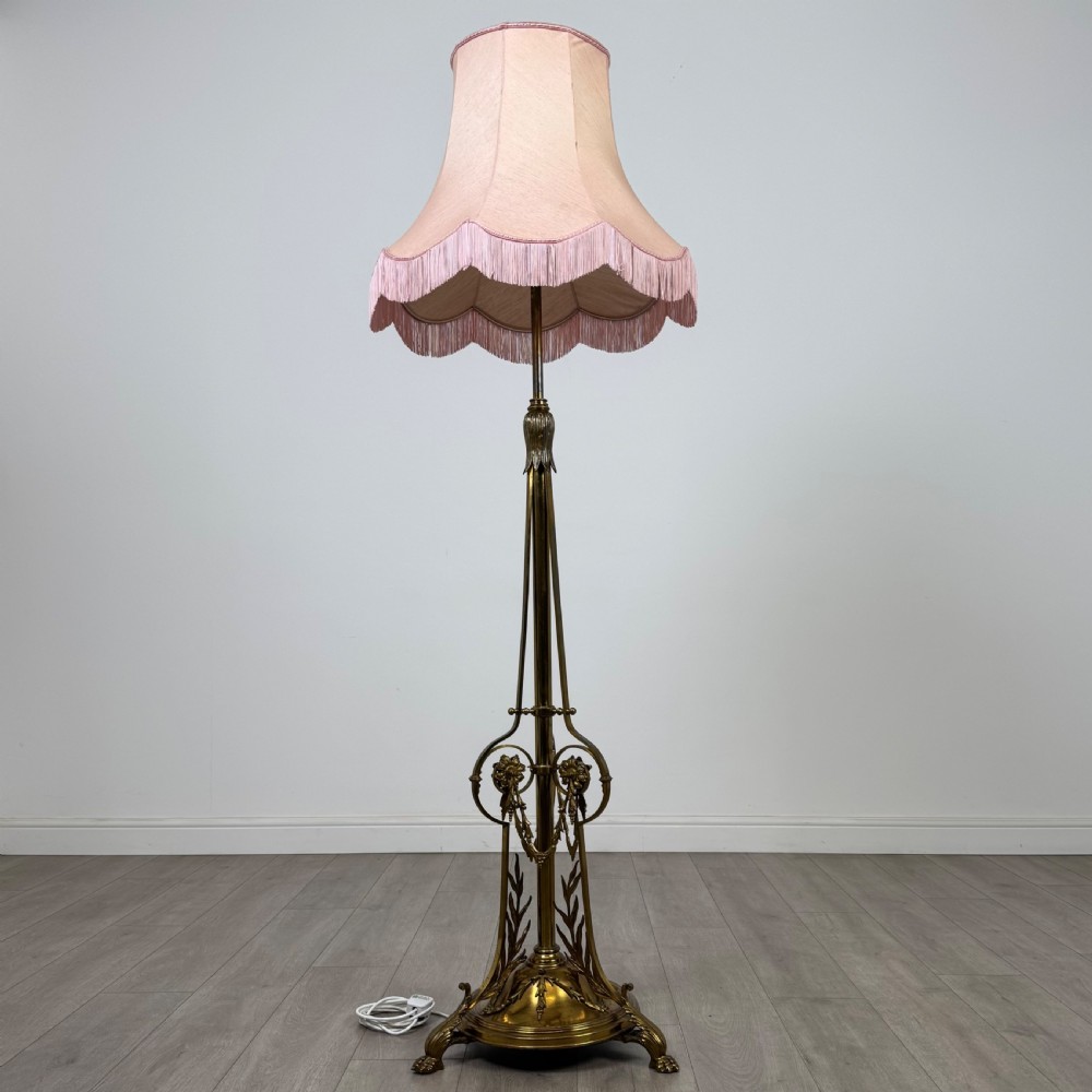 An antique 19th-century brass floor standing lamp featuring an ornate cast base with claw feet and a pink fringed shade.
