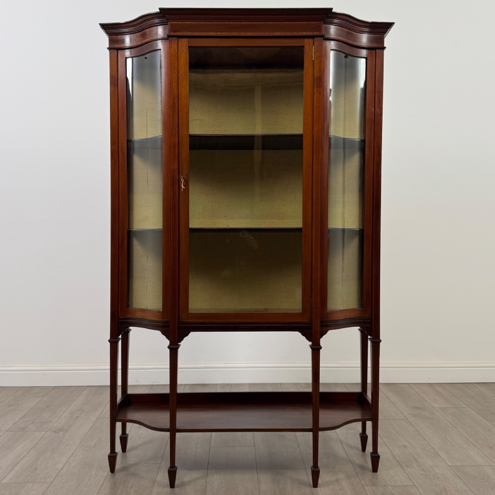 An Edwardian mahogany display cabinet featuring curved glass side panels, a glazed front door, fabric-lined interior shelves, and tapered square legs with a lower undertier.