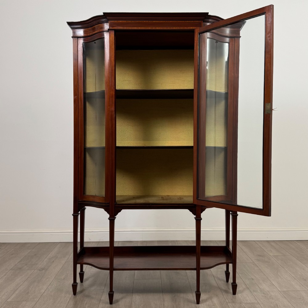 An Edwardian mahogany display cabinet featuring curved glass side panels, a glazed front door, fabric-lined interior shelves, and tapered square legs with a lower undertier. View II