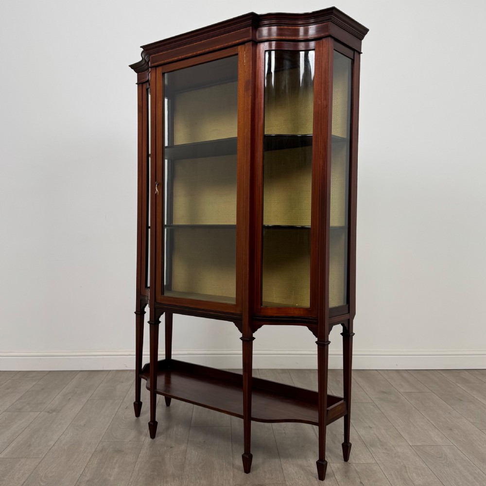 An Edwardian mahogany display cabinet featuring curved glass side panels, a glazed front door, fabric-lined interior shelves, and tapered square legs with a lower undertier. View III