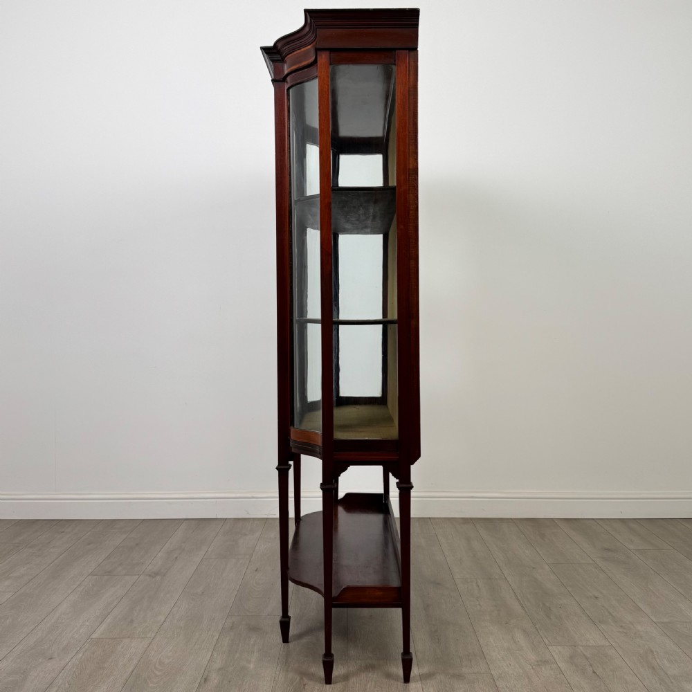 An Edwardian mahogany display cabinet featuring curved glass side panels, a glazed front door, fabric-lined interior shelves, and tapered square legs with a lower undertier. View IV