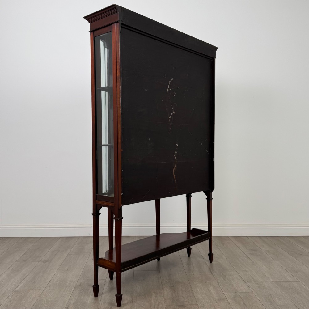 An Edwardian mahogany display cabinet featuring curved glass side panels, a glazed front door, fabric-lined interior shelves, and tapered square legs with a lower undertier. View V