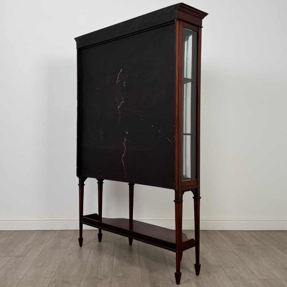 An Edwardian mahogany display cabinet featuring curved glass side panels, a glazed front door, fabric-lined interior shelves, and tapered square legs with a lower undertier. View VII
