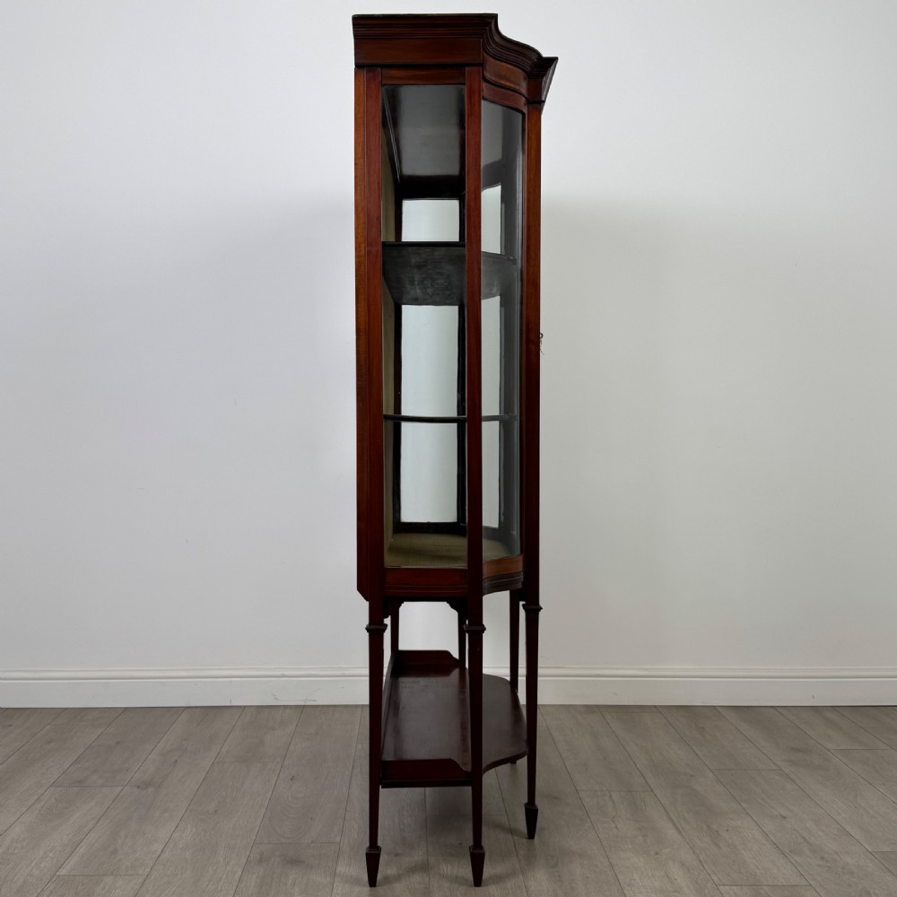 An Edwardian mahogany display cabinet featuring curved glass side panels, a glazed front door, fabric-lined interior shelves, and tapered square legs with a lower undertier. View VIII