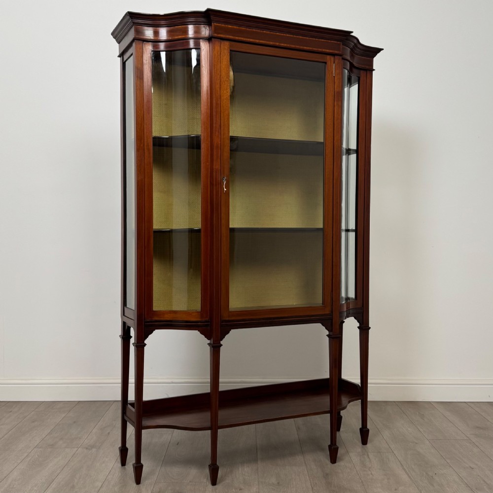 An Edwardian mahogany display cabinet featuring curved glass side panels, a glazed front door, fabric-lined interior shelves, and tapered square legs with a lower undertier. View IX