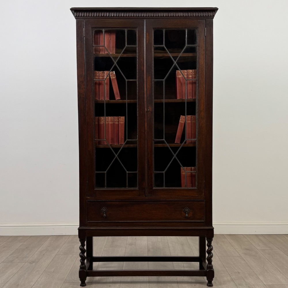 A dark oak Edwardian glazed display cabinet featuring two leaded glass doors, a lower drawer, and barley twist legs connected by stretchers.