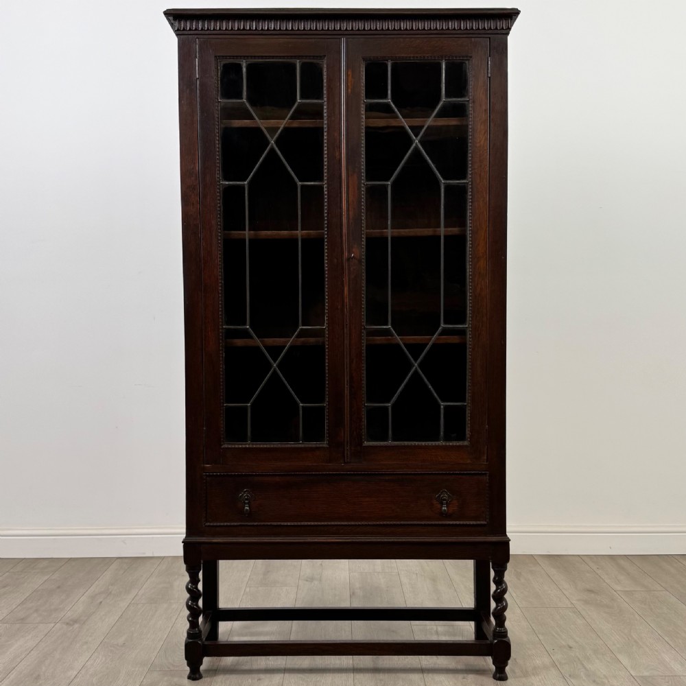 A dark oak Edwardian glazed display cabinet featuring two leaded glass doors, a lower drawer, and barley twist legs connected by stretchers. View II