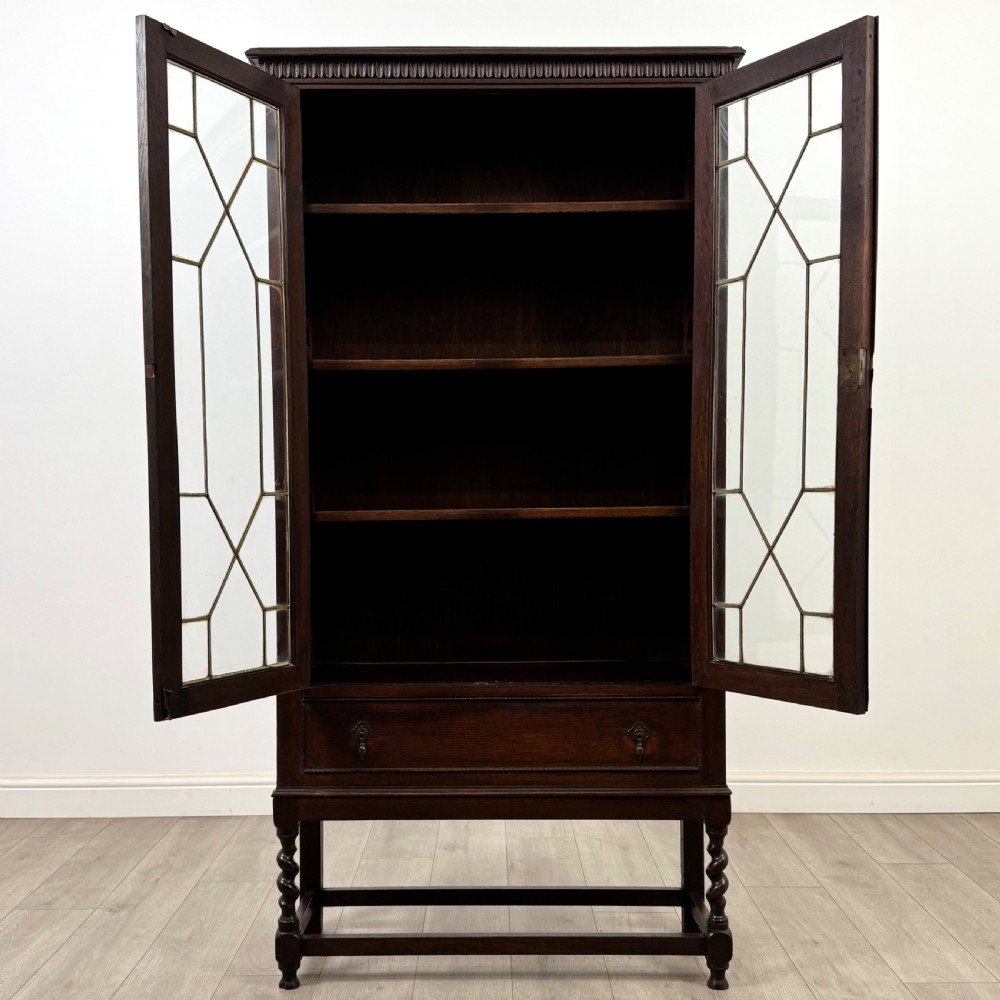 A dark oak Edwardian glazed display cabinet featuring two leaded glass doors, a lower drawer, and barley twist legs connected by stretchers. View III