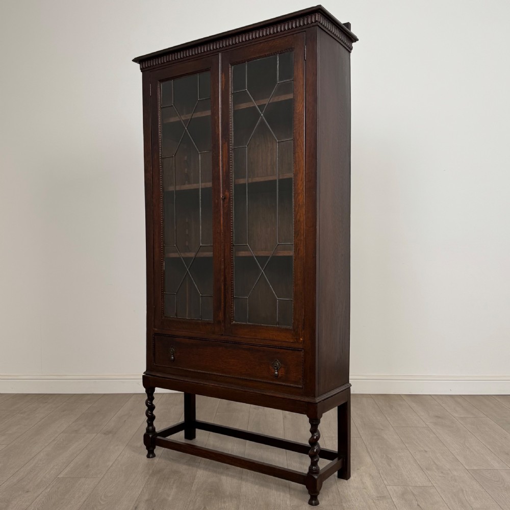 A dark oak Edwardian glazed display cabinet featuring two leaded glass doors, a lower drawer, and barley twist legs connected by stretchers. View IV