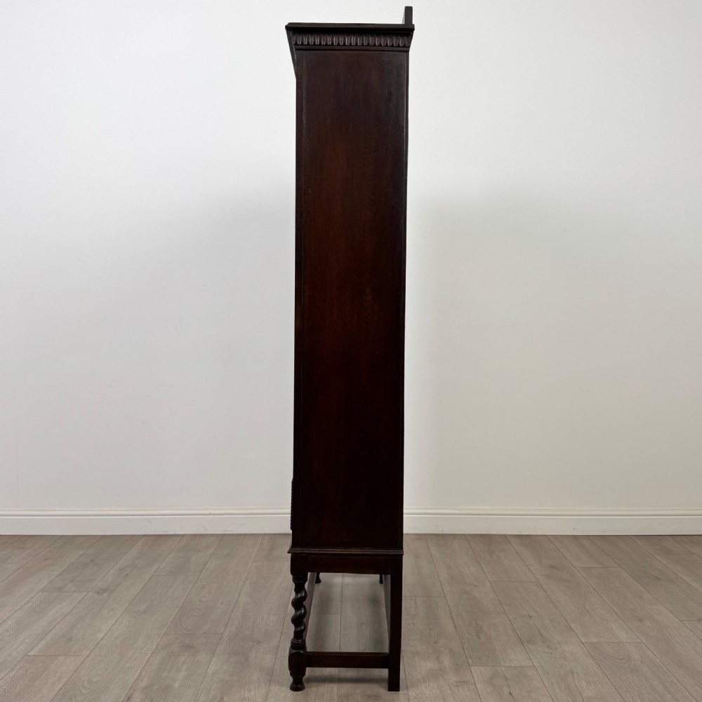 A dark oak Edwardian glazed display cabinet featuring two leaded glass doors, a lower drawer, and barley twist legs connected by stretchers. View V
