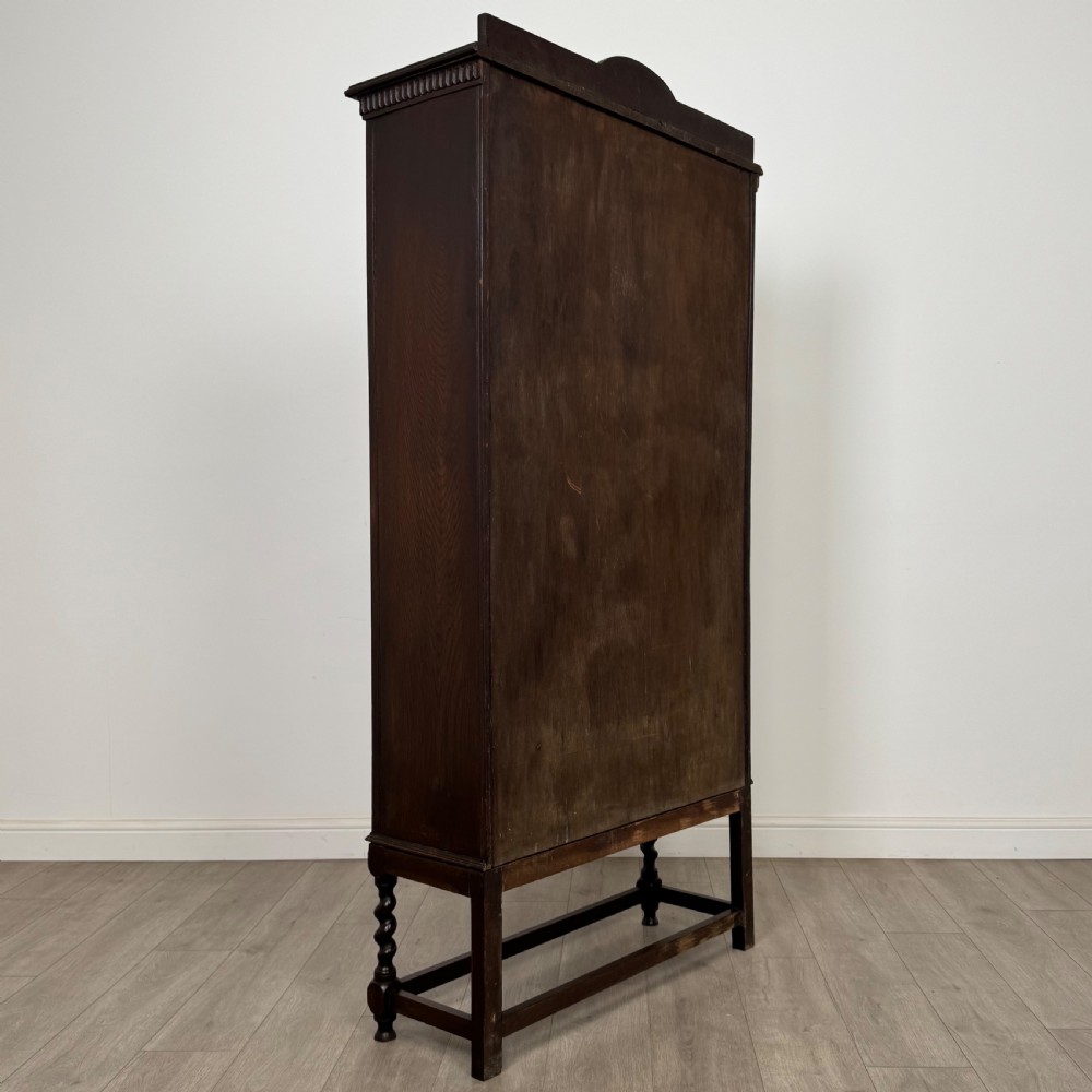 A dark oak Edwardian glazed display cabinet featuring two leaded glass doors, a lower drawer, and barley twist legs connected by stretchers. View VI