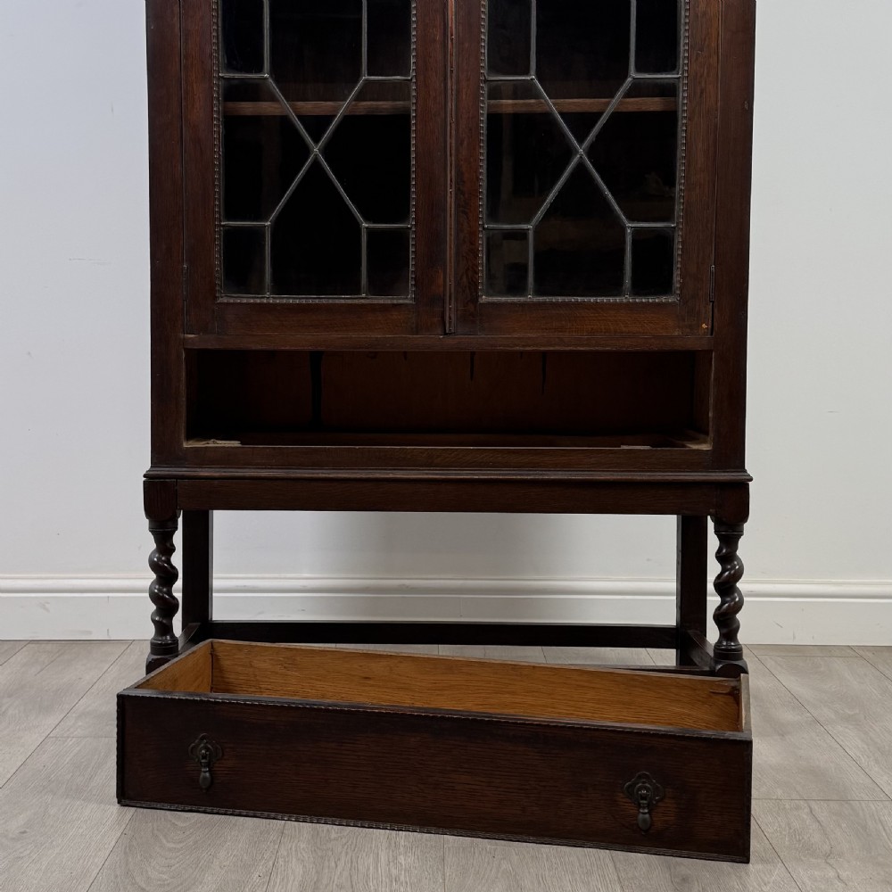 A dark oak Edwardian glazed display cabinet featuring two leaded glass doors, a lower drawer, and barley twist legs connected by stretchers. View IX