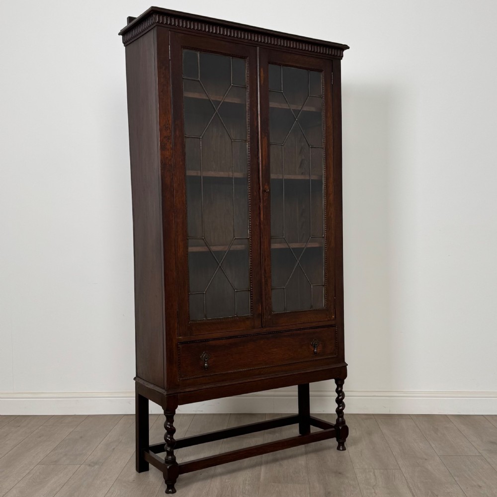 A dark oak Edwardian glazed display cabinet featuring two leaded glass doors, a lower drawer, and barley twist legs connected by stretchers. View XI