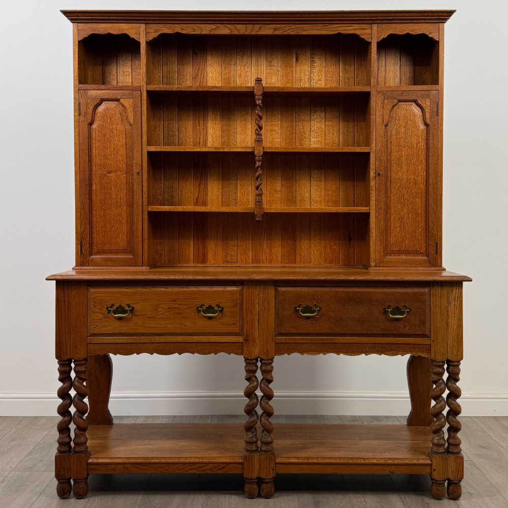 A golden oak kitchen dresser featuring an upper shelving rack with side cupboards and a lower base with two drawers, barley twist legs, and an open pot board.