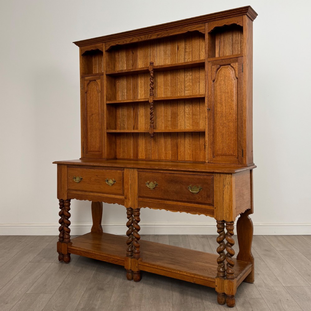 A golden oak kitchen dresser featuring an upper shelving rack with side cupboards and a lower base with two drawers, barley twist legs, and an open pot board. View III