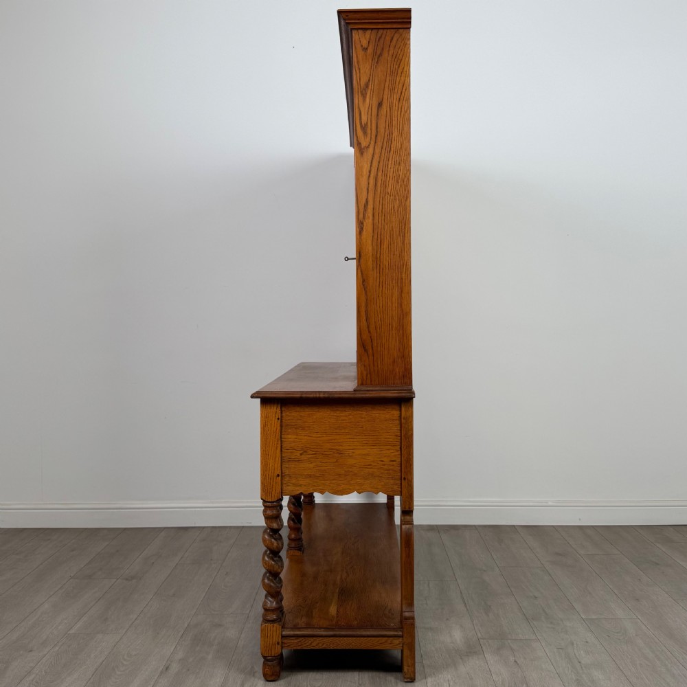 A golden oak kitchen dresser featuring an upper shelving rack with side cupboards and a lower base with two drawers, barley twist legs, and an open pot board. View IV