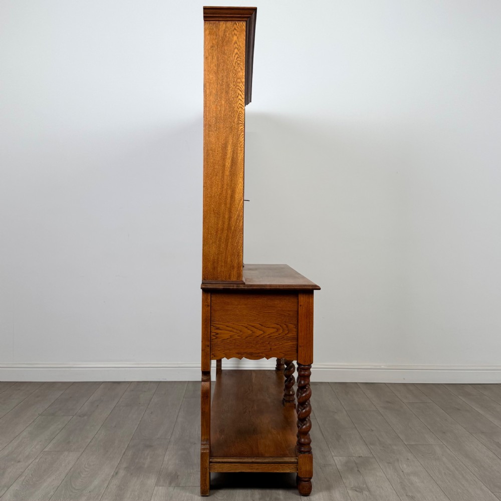 A golden oak kitchen dresser featuring an upper shelving rack with side cupboards and a lower base with two drawers, barley twist legs, and an open pot board. View VIII